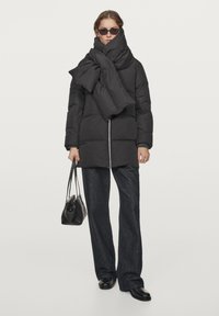 Zwarte pufferjack met een hoge kraag en een oversized sjaal, gecombineerd met donkere spijkerbroek en zwarte schoenen, met een kleine zwarte tas in de hand.