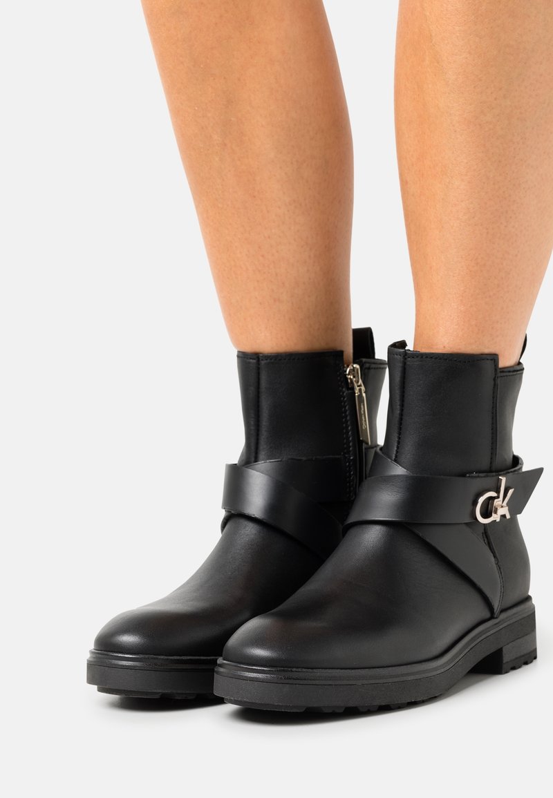 Botines negros de cuero suave, con cremallera lateral, una cinta ancha alrededor de la parte media del pie y un acento de logotipo plateado. Suela plana.