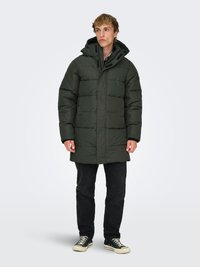 Parka imbottita verde con cappuccio, caratterizzata da un design trapuntato, chiusura con cerniera e dettagli neri. Indossata sopra jeans scuri e sneakers.