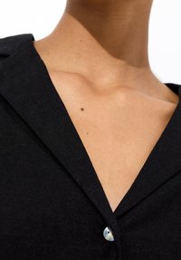 Camisa negra de lino con un escote en V profundo y cuello puntiagudo. Cierre de un solo botón con acabado brillante. Textura suave al contacto con la piel.