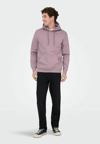 Jonge man die glimlacht en zijwaarts kijkt, gekleed in een mauve hoodie, zwarte jeans en zwarte sneakers, staand tegen een lichte achtergrond.