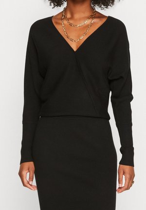 Kvinde iført en sort langærmet V-hals top og nederdel med lagdelte guldkæder og en ring på højre hånd.