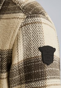 Beige en bruin geruit flanellen stof met een gestructureerde afwerking. Beslist met een zwart rubberen badge op de schouder met een arendgrafiek en tekst.