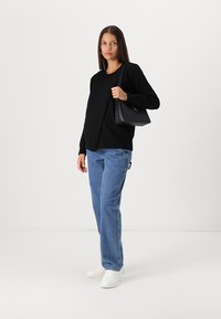 Pull en maille noir, jean bleu, baskets blanches. Le mannequin tient un sac à main noir de forme structurée avec une anse. Tenue simple et décontractée.