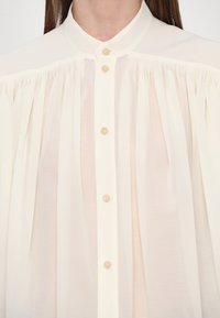 Svetlo béžová košeľa s gombíkmi a vysokým golierom, s prelínanou látkou pod ramennými švami a piatimi svetlými gombíkmi pozdĺž prednej strany.