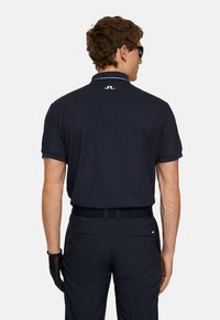 Marinblå pikétröja med korta ärmar, krage och vita accenter vid halsringningen, kombinerad med mörka byxor och en svart handske.