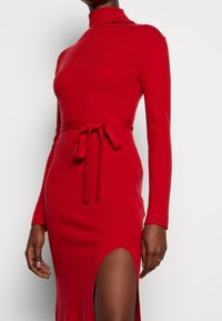Robe rouge à manches longues avec un col montant, dotée d'une ceinture nouée à la taille et d'une fente haute sur la cuisse, fabriquée dans un matériau doux et extensible.