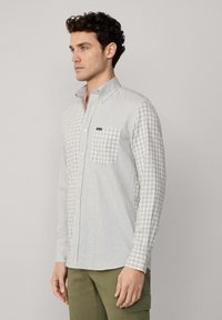 Camisa de botones en patrón de cuadros gris claro y blanco, de material de algodón, de manga larga, con cuello abotonado y que cuenta con un bolsillo en el pecho.