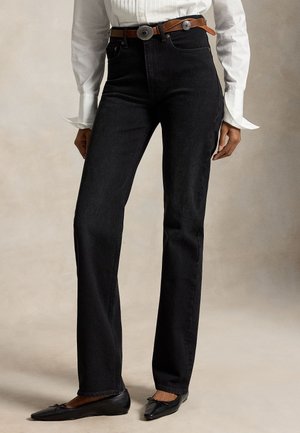 Femme portant un jean noir droit, une ceinture marron avec des conchos argentés, un chemisier blanc à manches longues et des ballerines noires, debout.