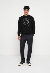 Felpa nera in cotone con una grafica che presenta un contorno bianco di un volto che indossa occhiali da sole. Abbinata a pantaloni scuri e sneakers nere.