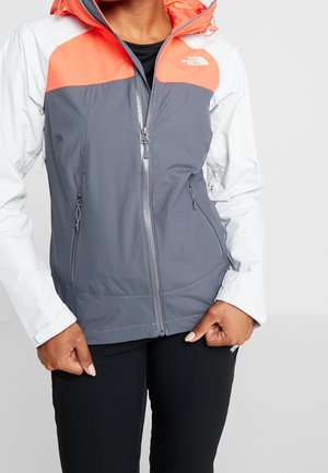 Persona con una chaqueta North Face con cremallera en gris, blanco y coral, y pantalones negros, de pie frente a un fondo claro y liso.