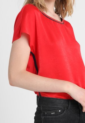 Kvinde iført en lys rød kortærmet bluse med sort kant, parret med sorte jeans, foran en ensfarvet lys baggrund.