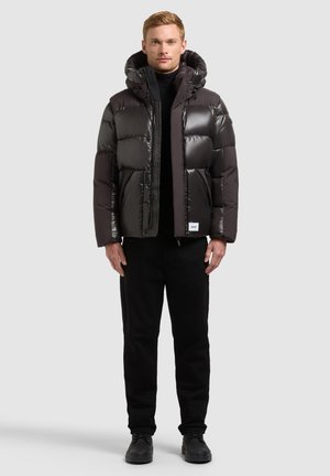 Homme portant une doudoune noire avec capuche, pantalon noir et chaussures noires, debout devant un fond clair uni.