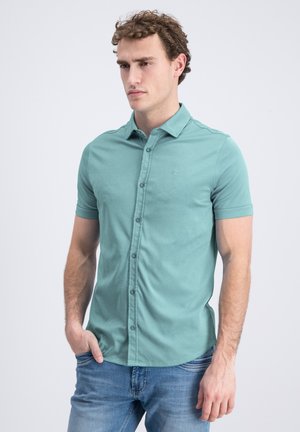 Jonge man in een teal gekleurde korte mouw blouse met knopen en een blauwe spijkerbroek, staand met één hand in de broekzak, neutrale uitdrukking, lichte achtergrond.