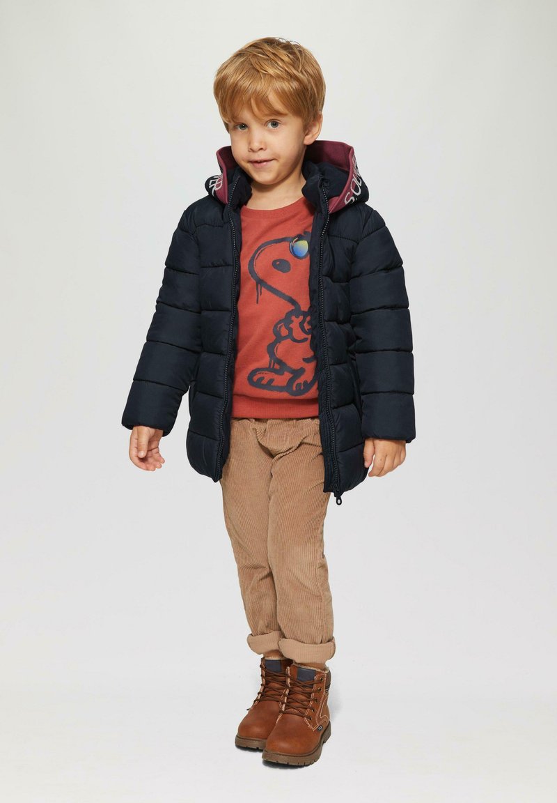 Marine pufferjack met bordeauxrode capuchon, oranje graphic sweater, beige corduroy broek enbruine boots met veters en rubberen zolen.