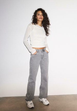 Žena s kučeravými vlasmi v bielom tričku s dlhým rukávom, sivých džínsach s širokými nohavicami a bielych teniskách, stojaca s rukami v vreckách.