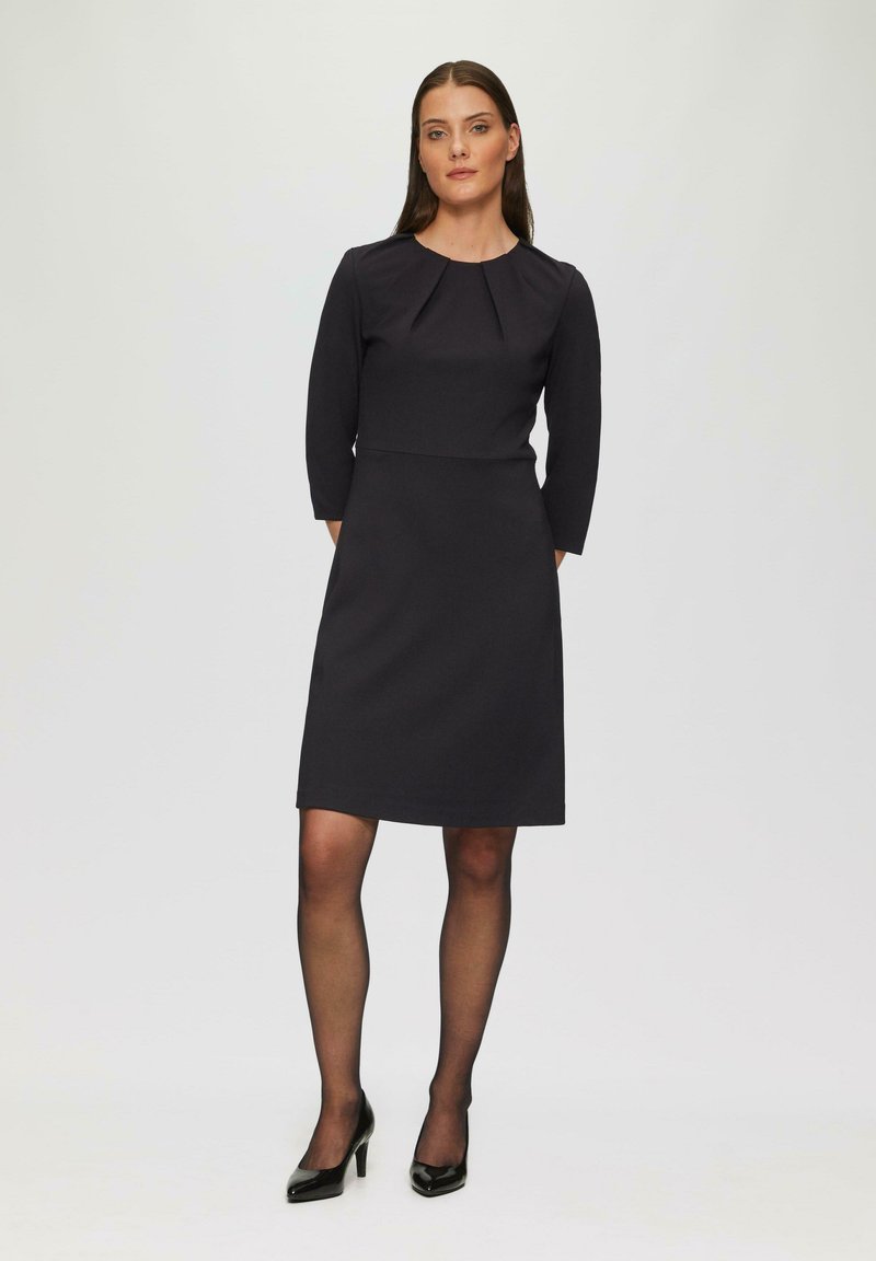 Robe noire ajustée avec des manches trois-quarts, encolure plissée et longueur au genou. Portée avec des collants noirs transparents et des talons noirs.