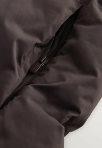 Prim-plan al unei jachete căptușite de culoare marron închis, parțial desfăcută, care arată textura țesăturii și detaliile fermoarului.