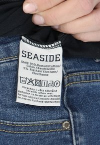 Etikette til denimtøj, hvidt stof med sort tekst. Indeholder plejeinstruktioner og materialeindhold: 95% bomuld, 5% elastan.