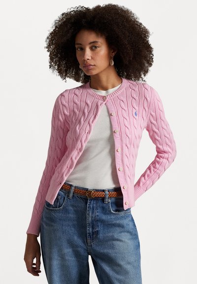 Woman with curly hair wears pink cable-knit cardigan over white shirt with blue jeans and brown braided belt, standing against white background.