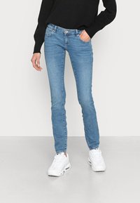 Jeans skinny azuis feitos de denim, com cinco bolsos, cintura com botão e costura subtil. Usados com ténis desportivos brancos.