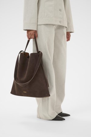 Personne portant un pantalon et une veste beige clair, des chaussures noires à bout pointu, tenant un grand sac fourre-tout en daim marron foncé avec deux poignées.