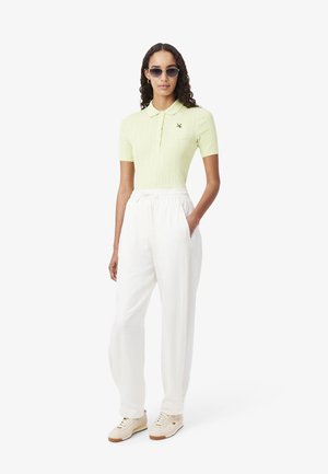 Femme portant un polo à manches courtes vert clair, un pantalon blanc à cordon, des baskets beiges et des lunettes de soleil, debout avec une main dans la poche.