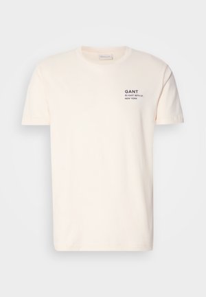 Krém színű pamut póló kerek nyakkal, rövid ujjakkal, az elején egy kis tengerészkék nyomattal, amelyen a "GANT 32 EAST 39TH ST. NEW YORK" felirat olvasható.