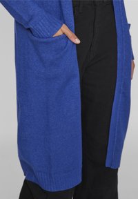 Largo cárdigan azul de suave tejido de punto, con un bolsillo frontal y puños acanalados, combinado con pantalones negros.