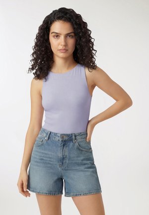 Donna con capelli ricci scuri che indossa una canotta lavanda senza maniche e pantaloncini di jeans a vita alta, in piedi con una mano sul fianco.