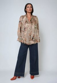 Bluse aus beigem, floral gemustertem Seidenstoff mit drapiertem Ausschnitt, kombiniert mit hoch taillierten, dunkelblauen weit geschnittenen Jeans und braunen Absatzschuhen.