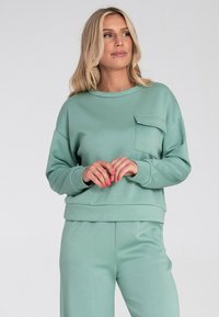 Miętowy sweter z okrągłym dekoltem i kieszenią z przodu, z prążkowanymi ściągaczami na mankietach i u dołu. Gładka tekstura i luźny krój.