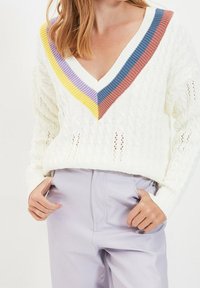 Pull en maille torsadée blanche avec un col en V profond, présentant des rayures côtelées multicolores en jaune, violet, bleu et rouille au niveau de l'encolure.