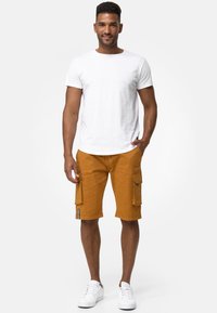 Camiseta blanca de cuello redondo combinada con pantalones cortos cargo mostaza a la altura de la rodilla, que cuentan con bolsillos laterales y un acento de logo, usados con zapatillas blancas.