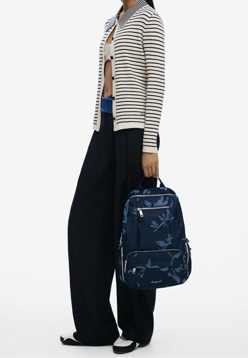 Personne portant un cardigan rayé et un pantalon large noir, tenant un sac à dos bleu marine à motif floral par la poignée supérieure.