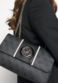 Černá kabelka s texturovanou kůží, zlatým řetízkovým popruhem, kulatým logem a růžovými proužky. Kompaktní a obdélníkový tvar.