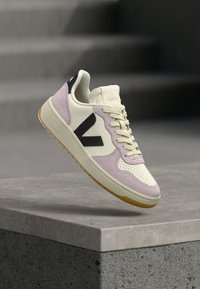 Αθλητικό παπούτσι με κρεμ επάνω μέρος, λιλά λεπτομέρειες, μαύρο λογότυπο και διάτρητη μύτη. Η σόλα από καουτσούκ προσφέρει αντίθεση με υφή φινιρίσματος.