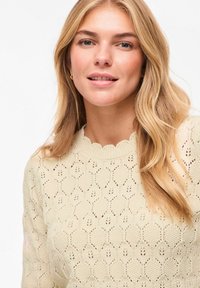 Jeune femme aux cheveux blonds longs portant un pull tricoté crème texturé avec un col festonné, souriant doucement sur un fond blanc.