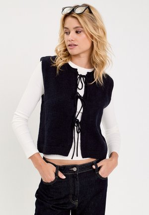 Marineblauwe gebreide vest met een ronde hals en strikjes aan de voorkant. Gedragen over een wit, langmouw shirt. Extra detail is de cropped fit.