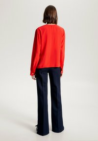 Blusa di seta rossa con vestibilità rilassata e maniche lunghe, abbinata a pantaloni blu navy su misura. L'outfit presenta una semplice chiusura a bottone ai polsini.