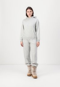 Hanorac gri din bumbac, cu o buzunar frontal și pantaloni de trening asortați. Model purtând cizme maro cu blană. Fundal neutru.