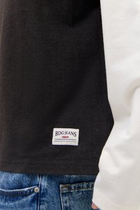 Κοντινή λήψη μαύρου πουκαμίσου BDG Jeans με λευκή ετικέτα πάνω σε μπλε τζιν και λευκό μανίκι ορατό στη δεξιά πλευρά.