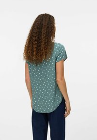 Kortærmet, teal bluse med hvide prikker, buet kant, let stof og løs pasform, parret med blå vide bukser.