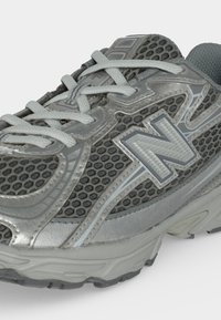 Κοντινή λήψη ενός γκρι και ασημί αθλητικού sneaker με δίχτυ, με λογότυπο "N" και ανοιχτογκρι κορδόνια σε λευκό φόντο.