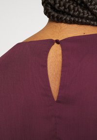 Lilla bluse med detalje, der har en nøglehulåbning på ryggen, afsluttet med en knaplukning og tekstureret stof.