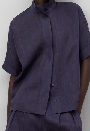 Persona con una camisa de manga corta abotonada, holgada y de color azul oscuro con sutiles rayas verticales, combinada con pantalones a juego.