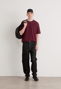 Bordeauxfarbendes T-Shirt, schwarze weite Jeans mit Taschen, schwarze Sneakers und eine schwarze Mütze. Das Model hält einen schwarzen Rucksack über einer Schulter.