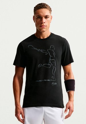 Homme portant un t-shirt noir Nike avec un dessin au trait d’un joueur de tennis frappant une raquette, un short blanc et un bracelet noir.