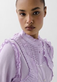 Lichtpaarse blouse met gerimpelde accenten, textuurstof en een knoopsluiting aan de voorkant met een hoge kraag.