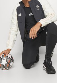 Schwarz-weiße Sportjacke mit cremefarbenen Ärmeln, kombiniert mit schwarzen Hosen und einem schwarzen Hemd. Ein Fußball liegt neben der Person auf dem Boden.